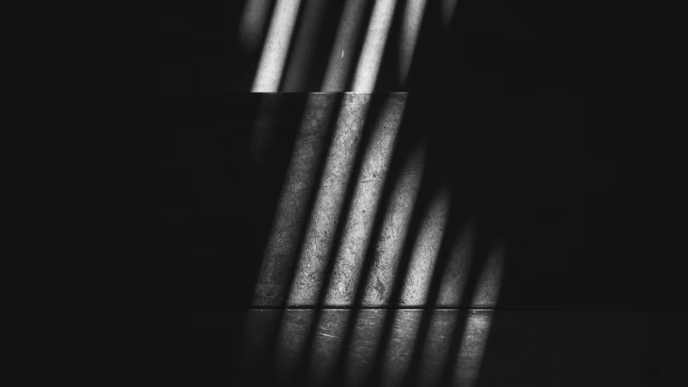 Striped light from a window blind falls across a dark stairwell wall in a stark abstract pattern.